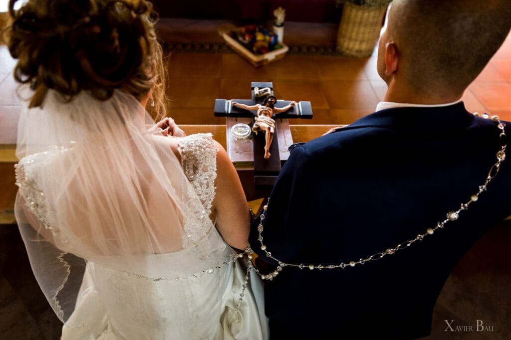 Close-up from behind of a bride in a veil and groom in a dark suit looking at a crucifix during a Catholic wedding ceremony or blessing. Rosary draped around the groom's shoulders.
