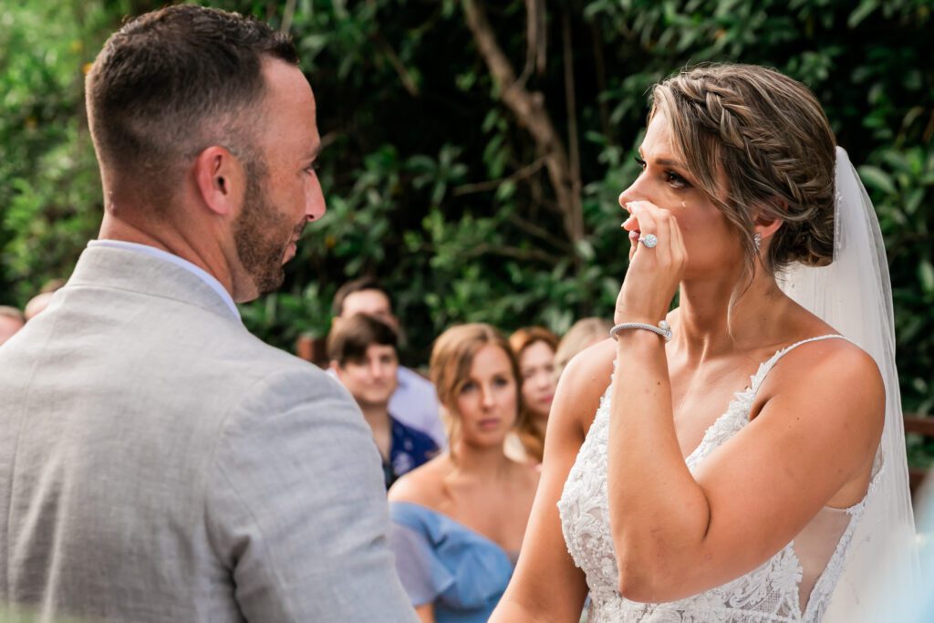 Emotional bride wiping a tear away while looking at the groom during the wedding vows, with the bridal party standing in the background. cancun wedding