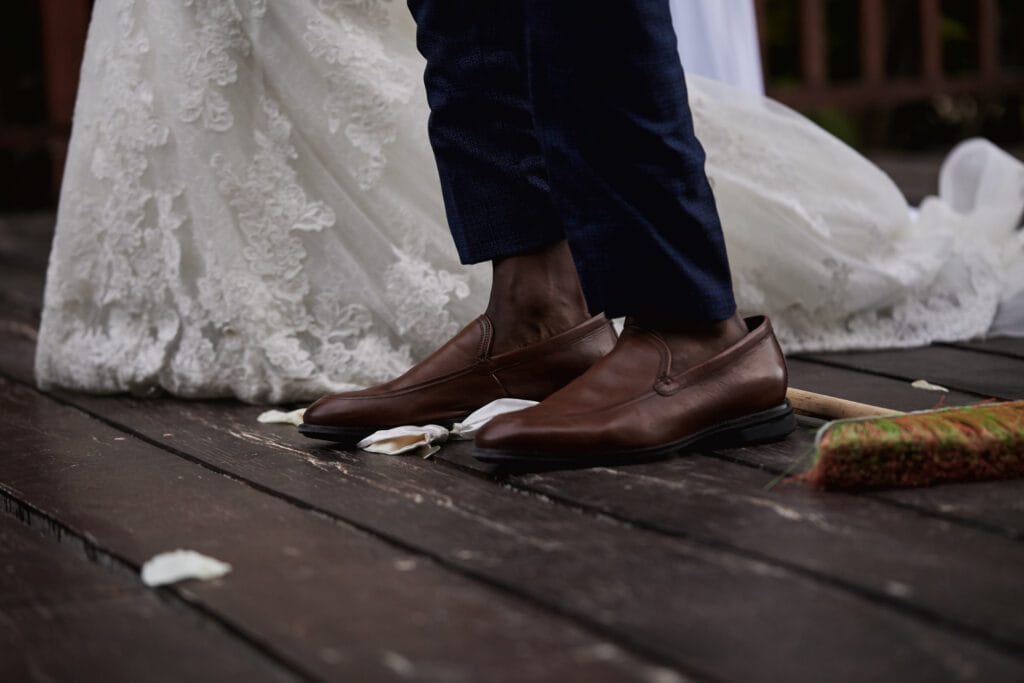 Close-up of the groom's brown leather shoes and the hem of the bride's white lace dress next to a decorated broom resting on the wooden floor, symbolizing the 'Jumping the Broom' unity ritual at a wedding.