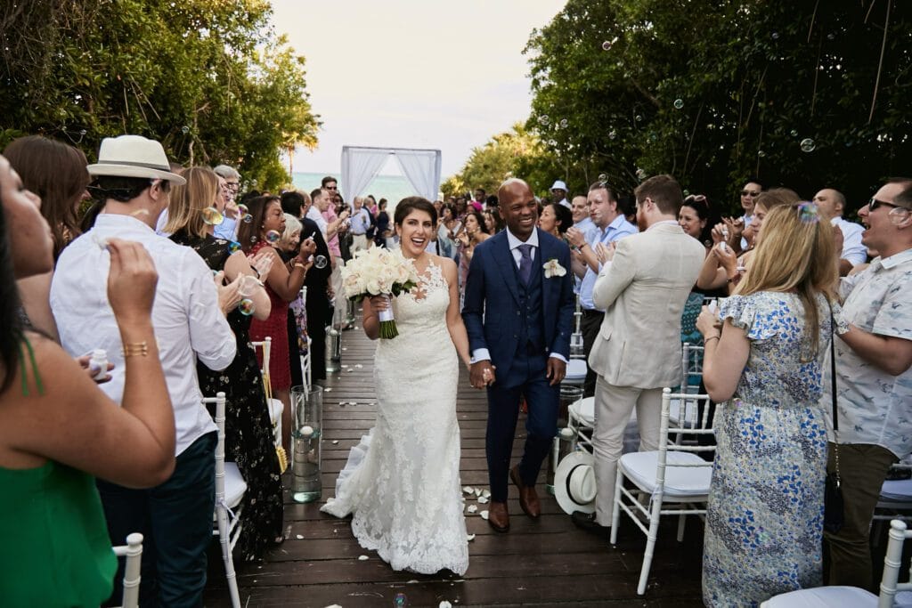 Happy newlywed couple, a bride in a lace dress and groom in a blue suit, smiling while walking down a wooden aisle surrounded by cheering guests at an outdoor tropical wedding ceremony.