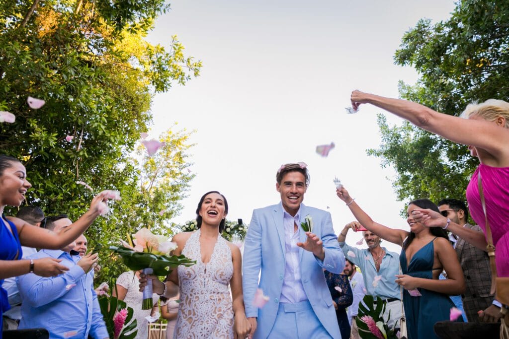 Excited newlyweds in tropical attire smiling as guests toss flower petals at them during a joyous wedding recessional at a Cancun destination wedding.