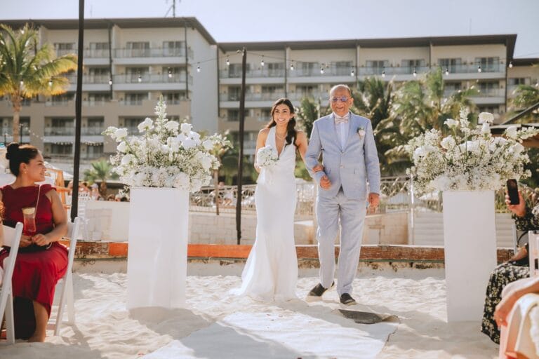 Kishory arriving at the beach ceremony between arrangements of white flowers.