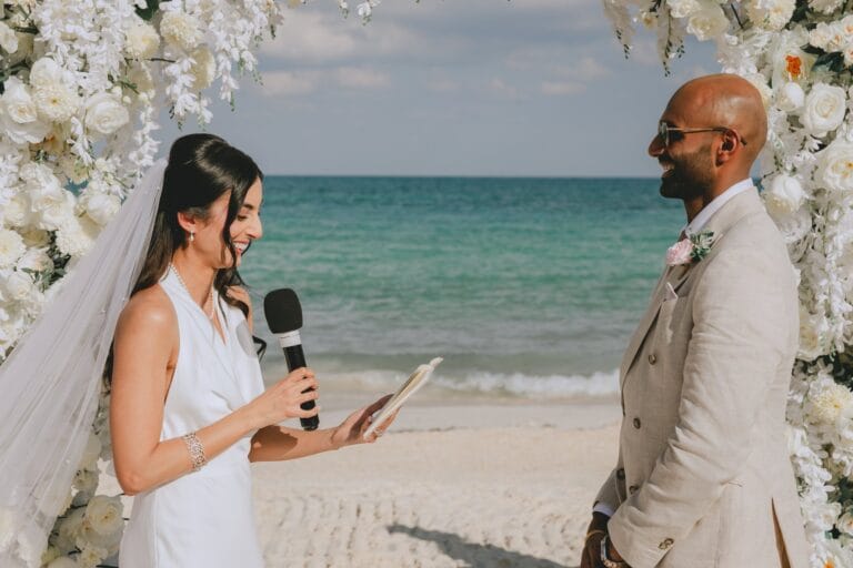 Kishory reading her vows to Kyle beneath a white floral arch on the beach.
