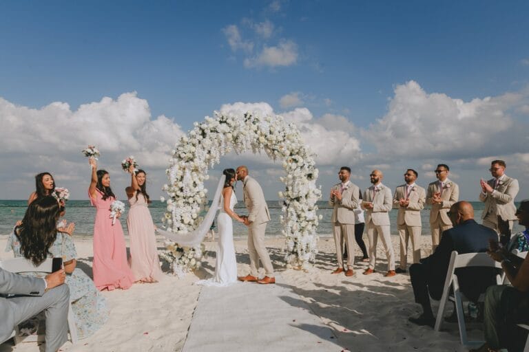 Kyle and Kishory kissing beneath their beach ceremony arch as guests watch.