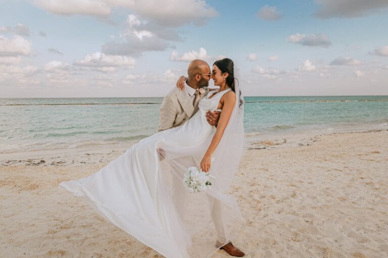 Kyle carrying Kishory in her white wedding dress beside the turquoise water.