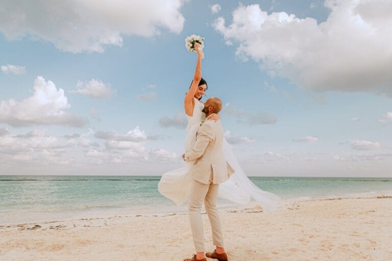 Kishory raising her bouquet while Kyle lifts her on the beach.