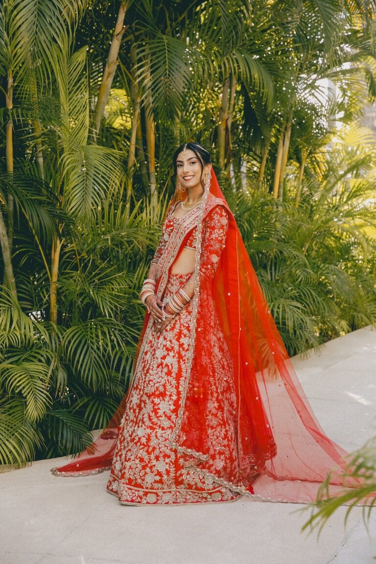 Full-length portrait of Kishory in her red lehenga beside tropical greenery.