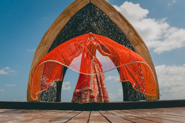 Kishory lifting her red dupatta in front of a pointed wooden arch.