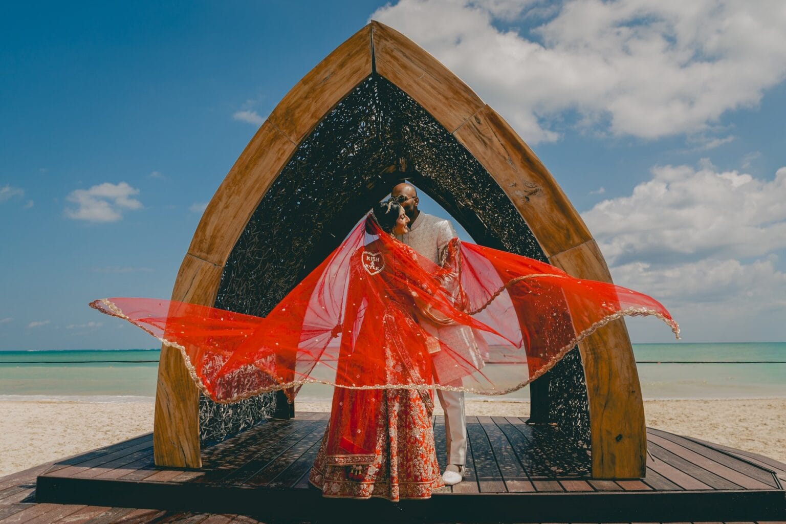 Kyle and Kishory embracing beneath a wooden arch as her red dupatta moves in the breeze.