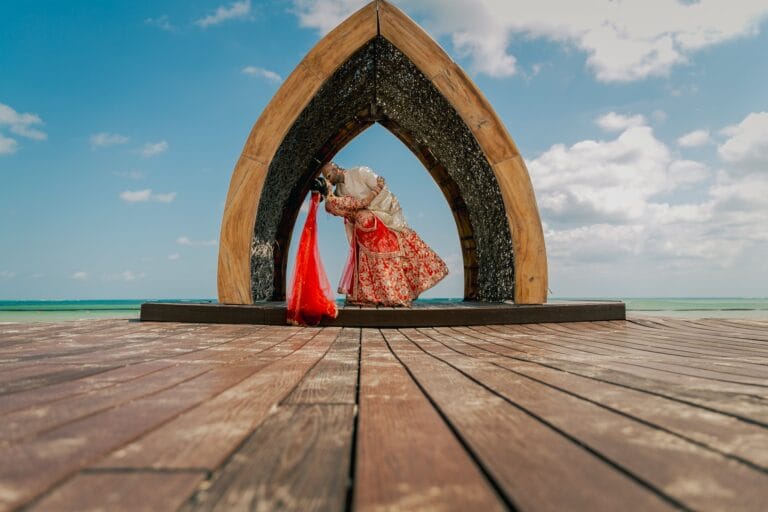 A wide portrait of Kyle and Kishory beneath the wooden arch on the beachfront deck.