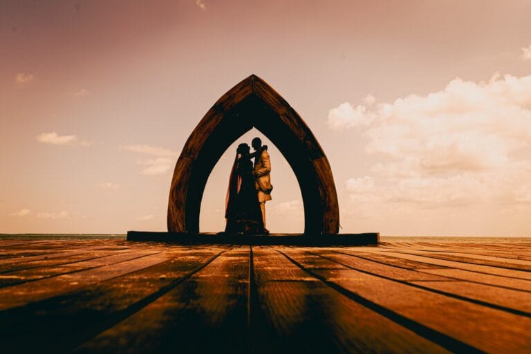 Kyle and Kishory silhouetted beneath the wooden arch against a warm sunset sky.