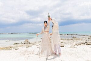 Nusaibah and Jai raising a bottle together beside the sea in their wedding attire.